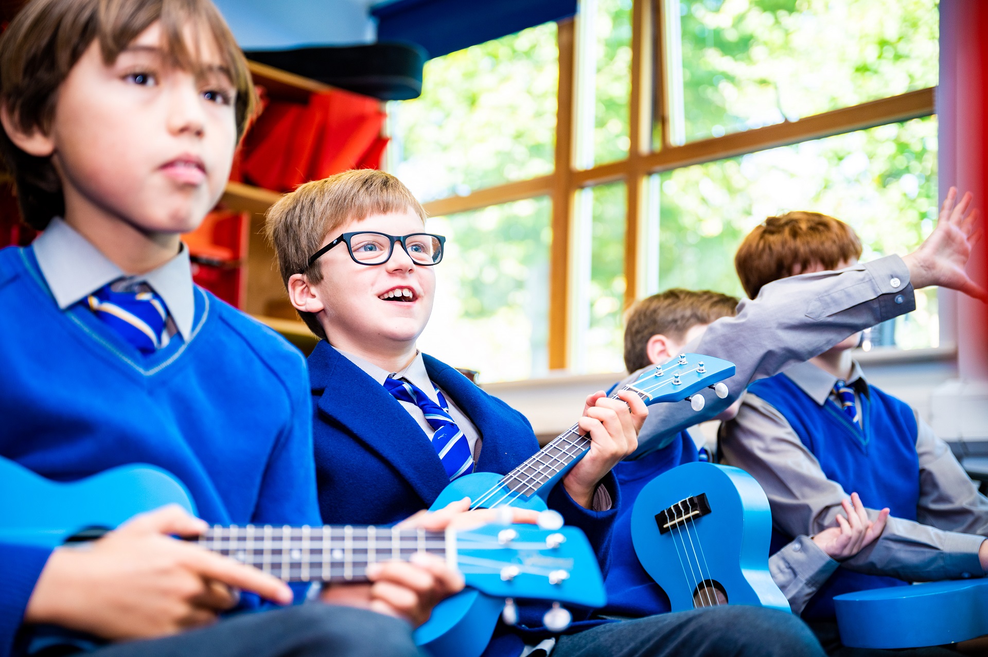 Pupils sit in a music lesson wearing a blue school uniform. They are playing on guitars. The child in focus in the image has brown hair and is wearing glasses, smiling and looking at the teacher.