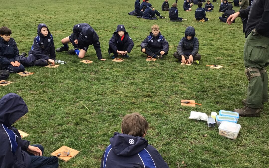 A group of school pupils in blue PE kit sit in circles on the grass in a town square. There are houses and a line of trees in the background.