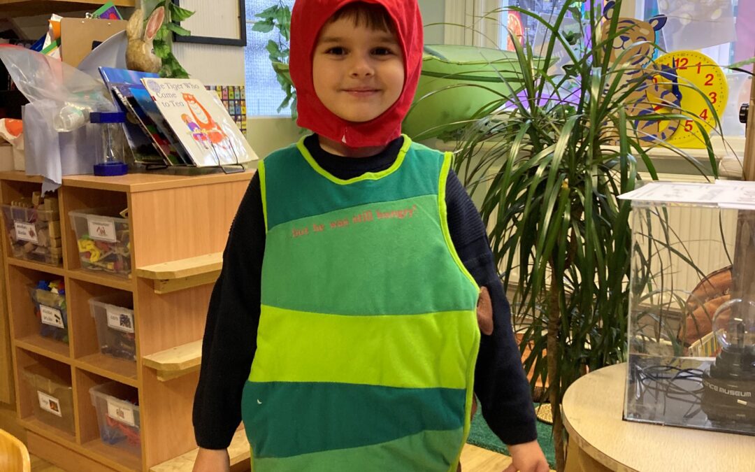 A boy stands in his classroom dressed up in a green caterpillar costume with a red hat on.