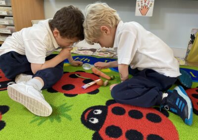 Two boys wearing a white top and navy tracksuit bottoms sit on a carpet in a classroom. They are looking into magnifying classes to see objects on the floor.