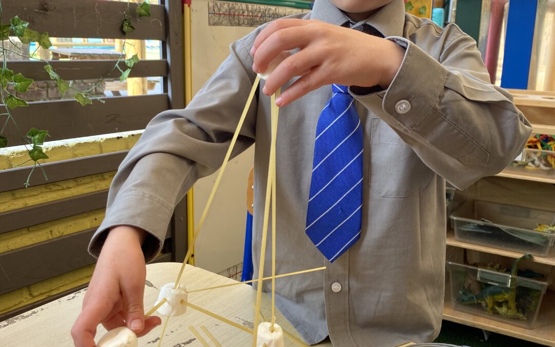 A boy in a blue school uniform is outside at school - he is holding up a model he has made out of spaghetti and marshmallows.