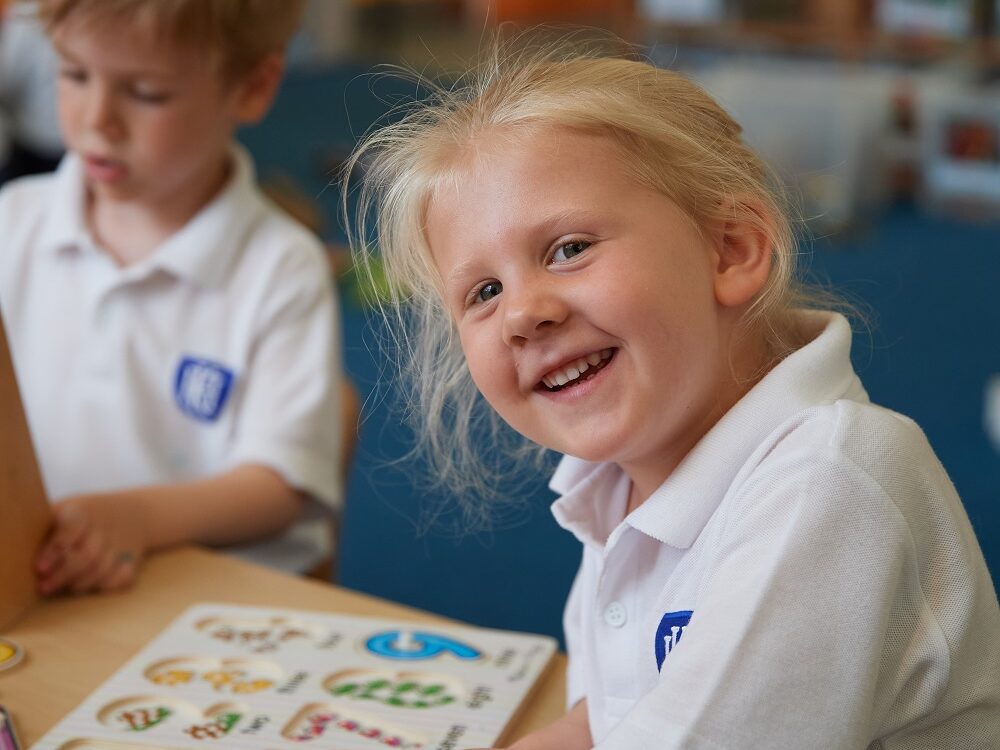 A Nursery child sits at a desk in Caterpillar classroom, smiling. She is wearing a white polo top.
