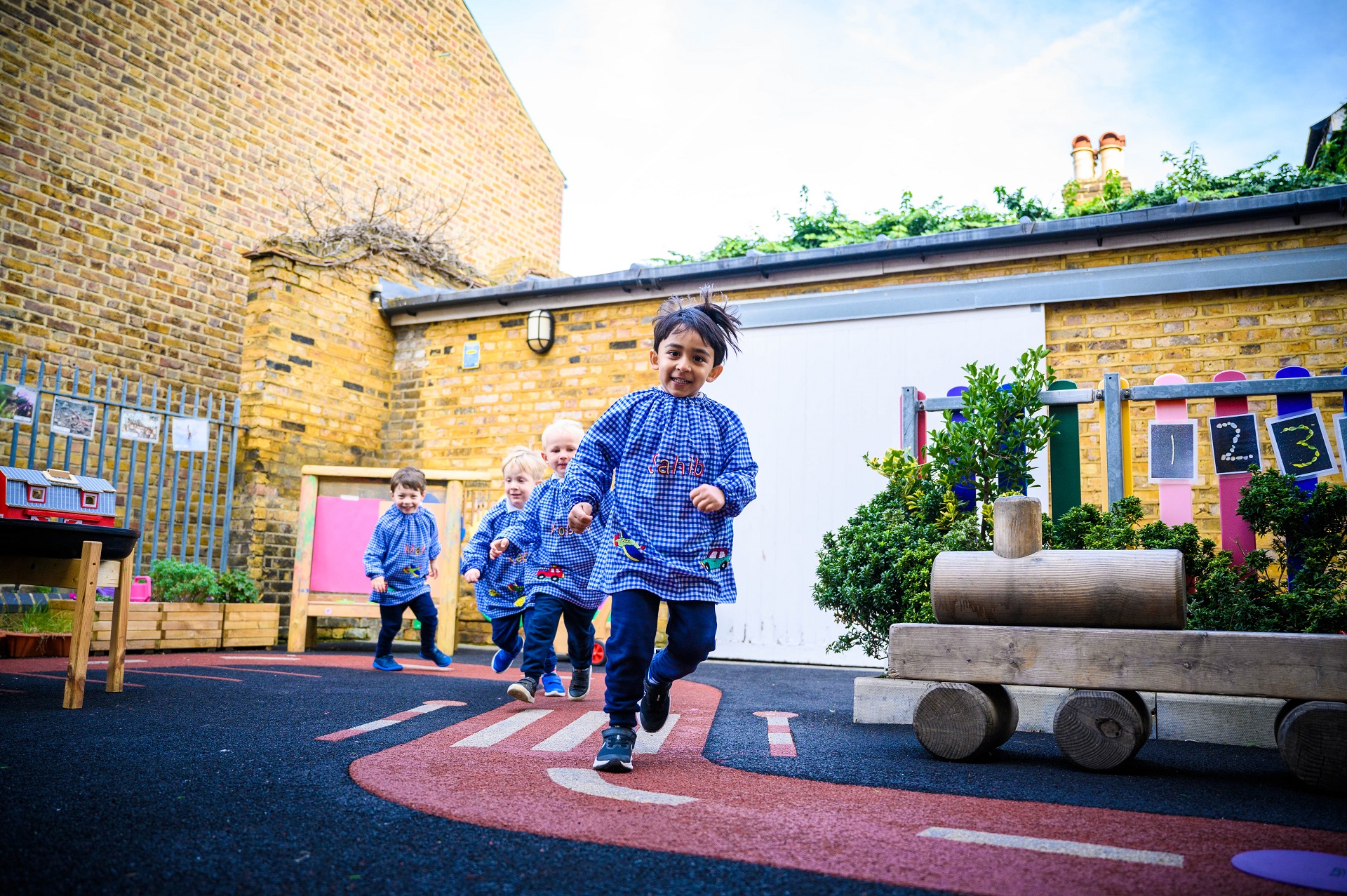 Four Nursery pupils run across the playground wearing the blue gingham KHN smocks.